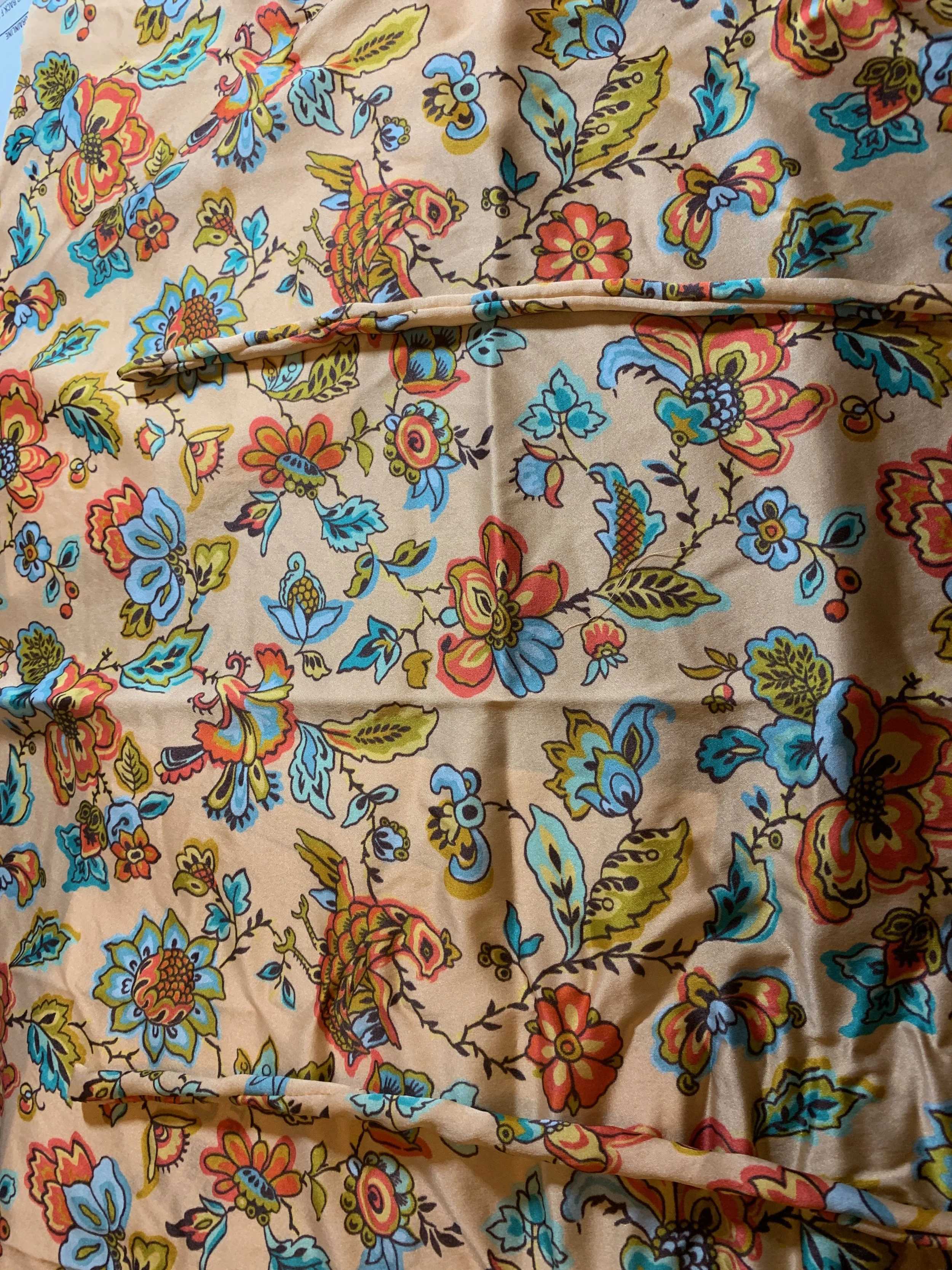 Close-up of a beige fabric with a vivid floral and bird print in orange, blue, and green tones, folded with a sewn seam.