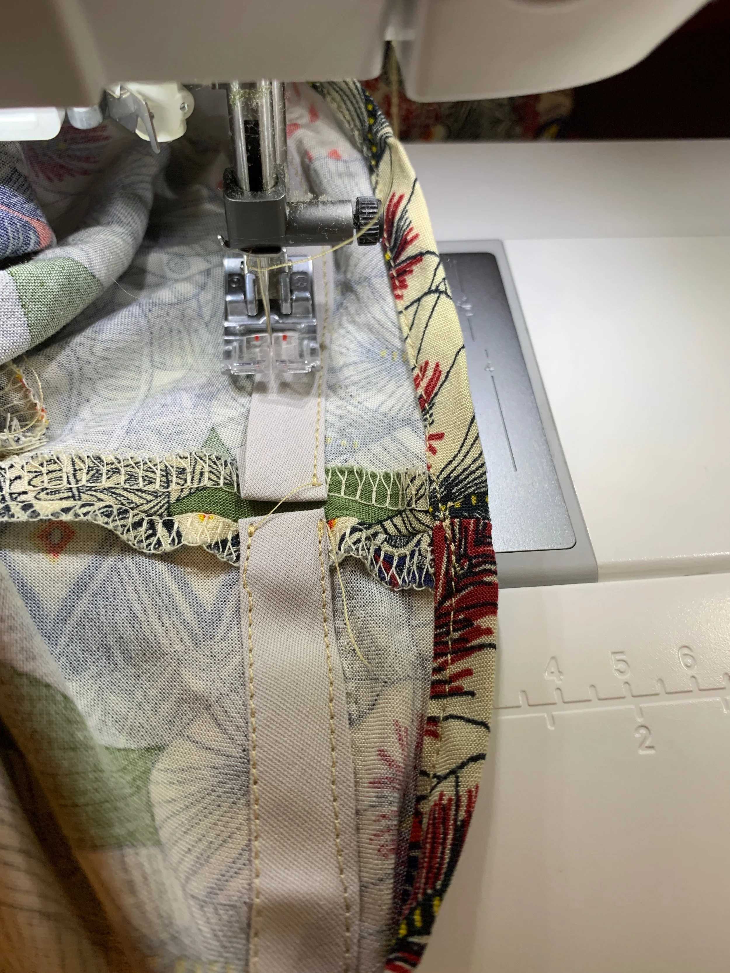 Close-up of patterned fabric being sewn with bias tape under a sewing machine foot, in cream, red, and green tones.