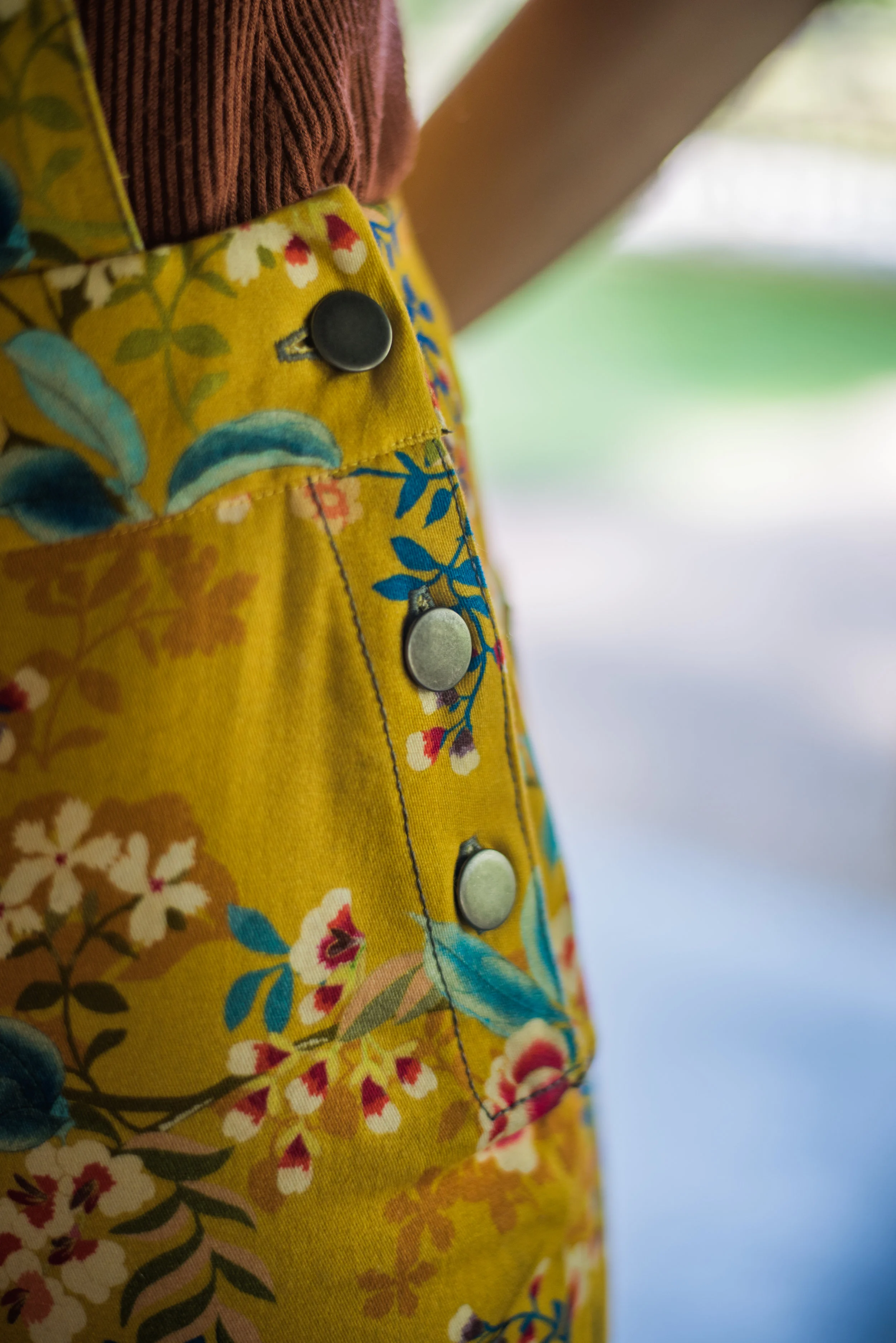 Close-up of yellow floral overalls fabric with metal snap buttons, set against a blurred green background.